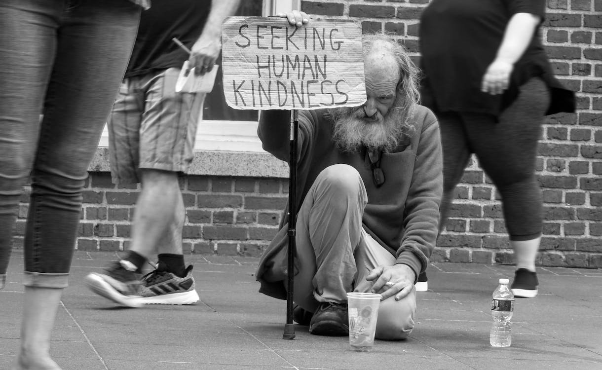 Man in Gray Sweater Sitting on Ground Holding A Sign