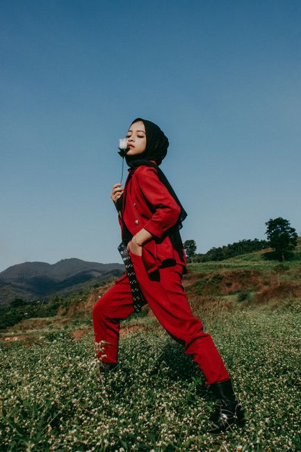 Portrait of a woman in a red outfit and hijab posing in a green field under a blue sky.
