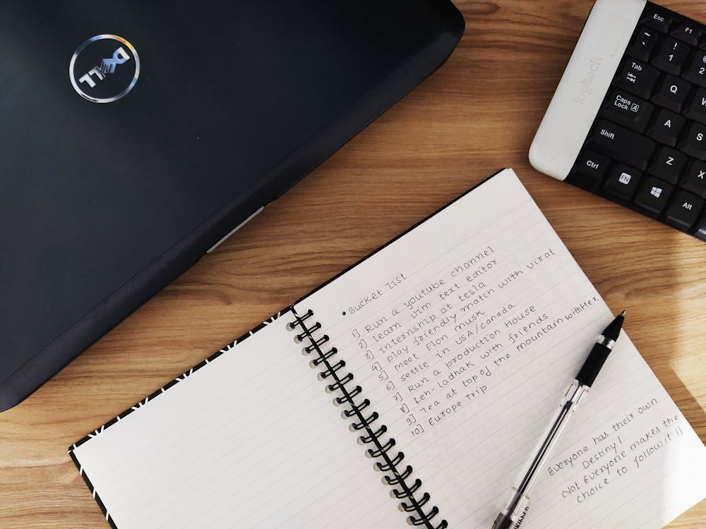 A Pen On Notebook With A Written Bucket List Beside A Laptop and Keyboard