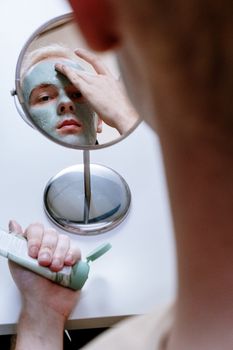 A man applies a green clay mask while looking into a mirror, emphasizing skincare routine.