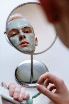 A man applying a green clay mask as part of his skincare routine reflected in a mirror.