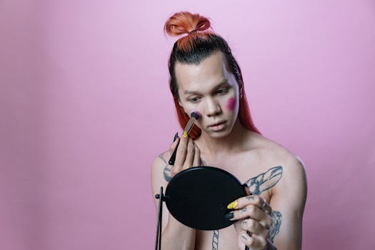 Man with hair bun applying makeup with a brush in a pink background studio setting.