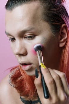 Close-up of a man applying blush with a brush against a pink backdrop, highlighting expressive makeup artistry.