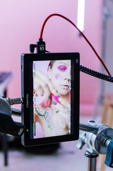 A makeup artist applies vibrant colors to a model on a screen, set against a pink background.