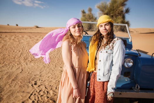 Two women in colorful attire posing by a vintage vehicle in the desert, enjoying a sunny day.