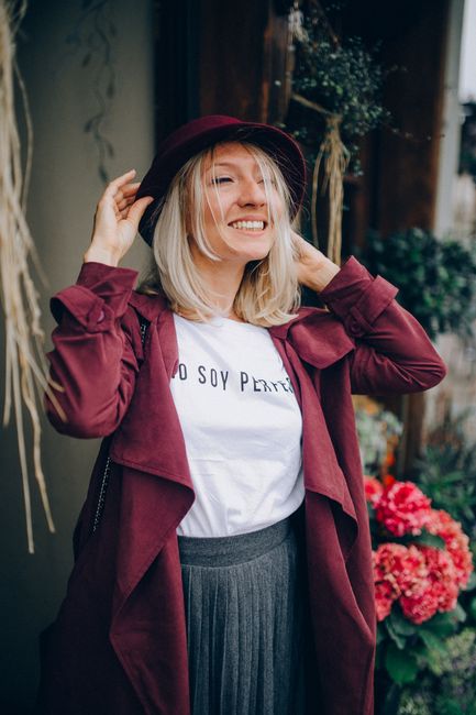 Fotomural Mujer sonriente con un atuendo burdeos elegante ajustándose el sombrero, de pie junto a flores vibrantes.