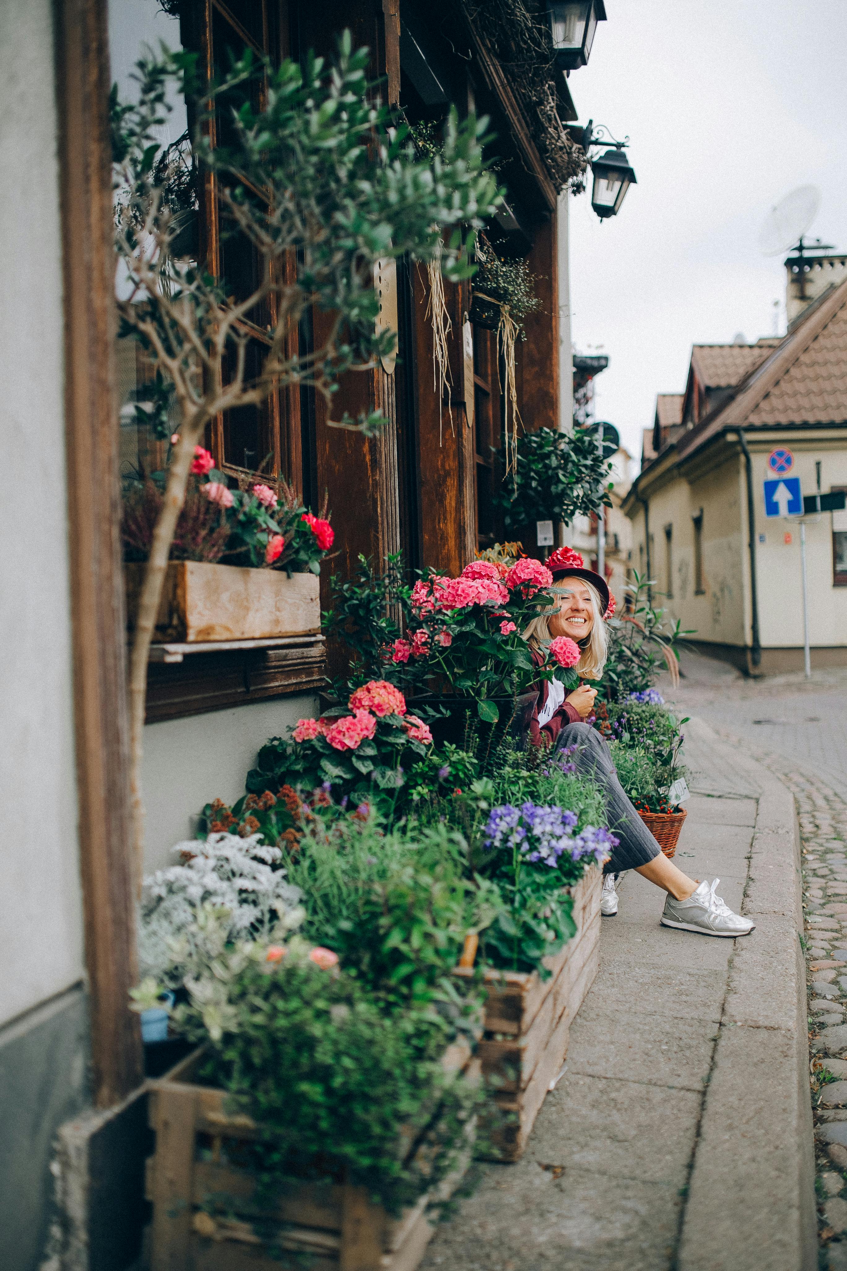 Free A woman enjoys a peaceful moment sitting among blooming flowers on a picturesque, plant-lined street. Stock Photo