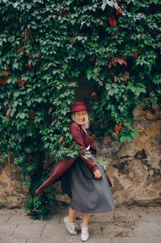 Stylish adult woman in autumn attire posing by a lush green ivy wall, exuding elegance and joy.