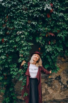 Caucasian woman with blond hair smiling in a hat and coat against a leafy wall.