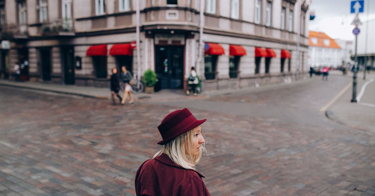 Blond woman in a red coat and hat walks down a cobblestone street in a European city.