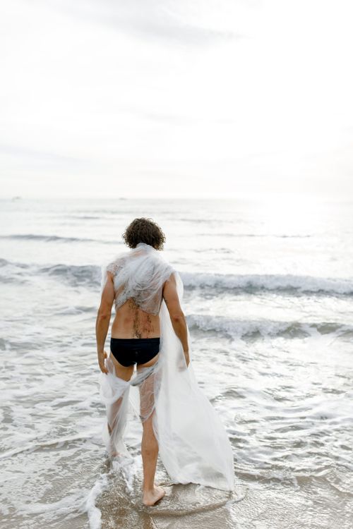 Free Back view of a shirtless man wrapped in plastic sheet standing on a sandy beach by the ocean waves. Stock Photo