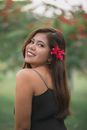 Side view attractive content ethnic female decorating long hair with red flower and looking at camera over shoulder while standing in lush garden