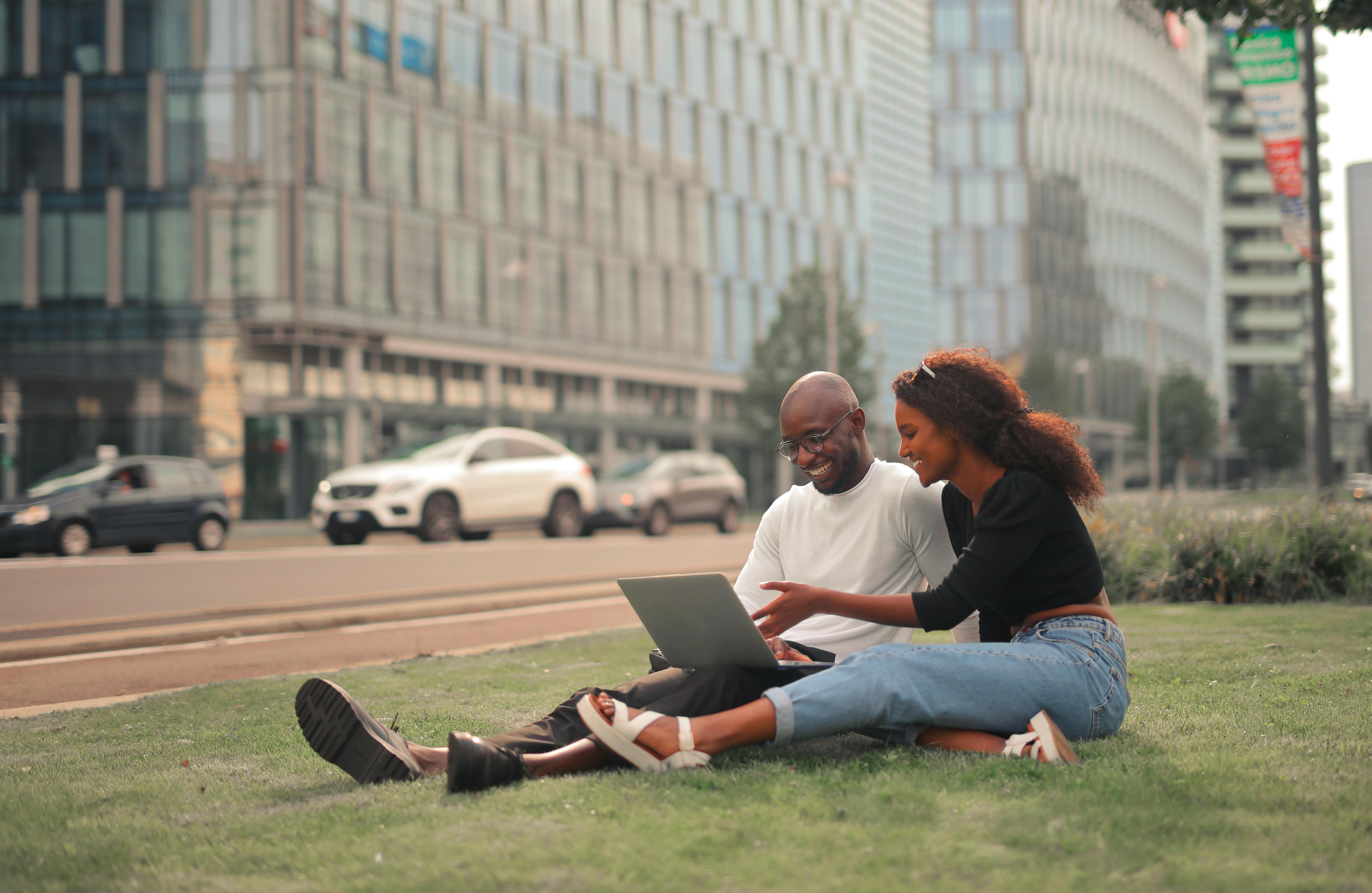 Young couple enjoying modern technology while sitting on grass in Milan's urban landscape.