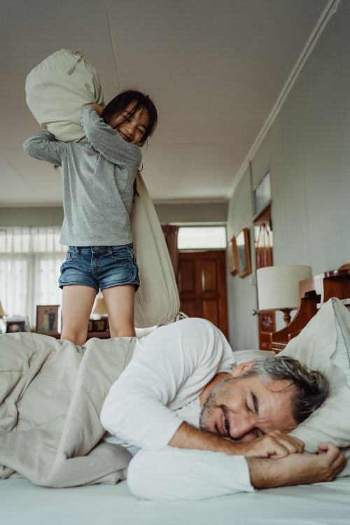 Free A joyful pillow fight between a father and daughter in a comfortable, light-filled bedroom. Stock Photo