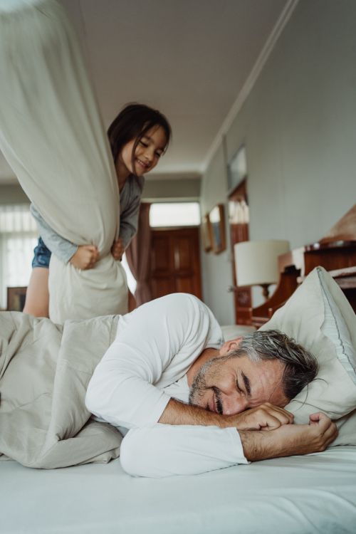 Free Playful father and daughter having fun with a pillow fight in a cozy bedroom setting. Stock Photo