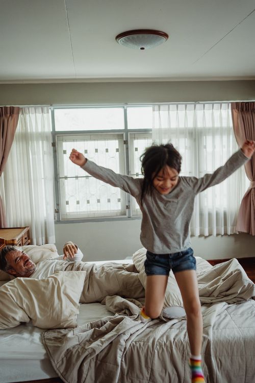 Free Child joyfully jumping on a bed with a smiling parent in a cozy bedroom setting. Stock Photo