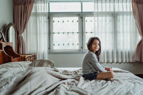 Free A young girl sitting on a bed in a bright, cozy bedroom with natural lighting. Stock Photo