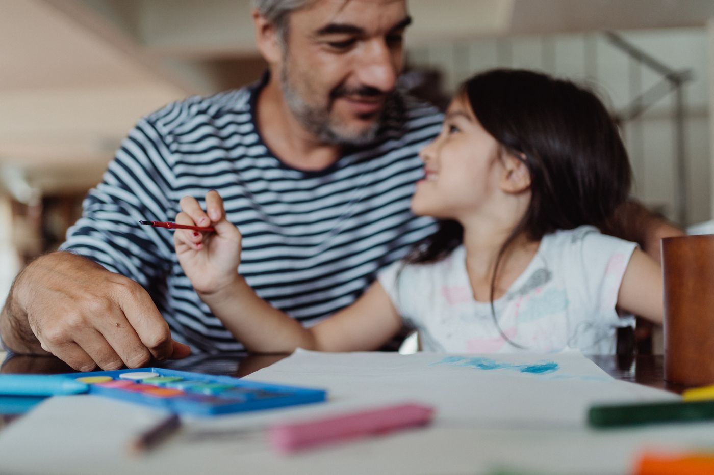 A father and his young daughter drawing and painting together at a desk at home.