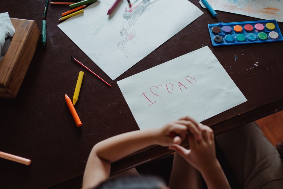 A child's handwritten note 'I ❤️ Dad' on paper surrounded by art supplies on a wooden table.