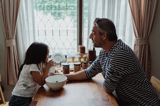 Father and daughter share a warm breakfast moment at home, sitting by a window.