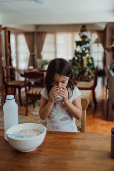 A girl drinks milk beside a cereal bowl in a festive indoor setting.