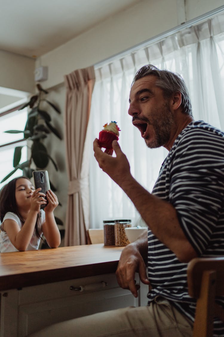 Girl Taking A Photo Of Her Father Eating A Cupcake