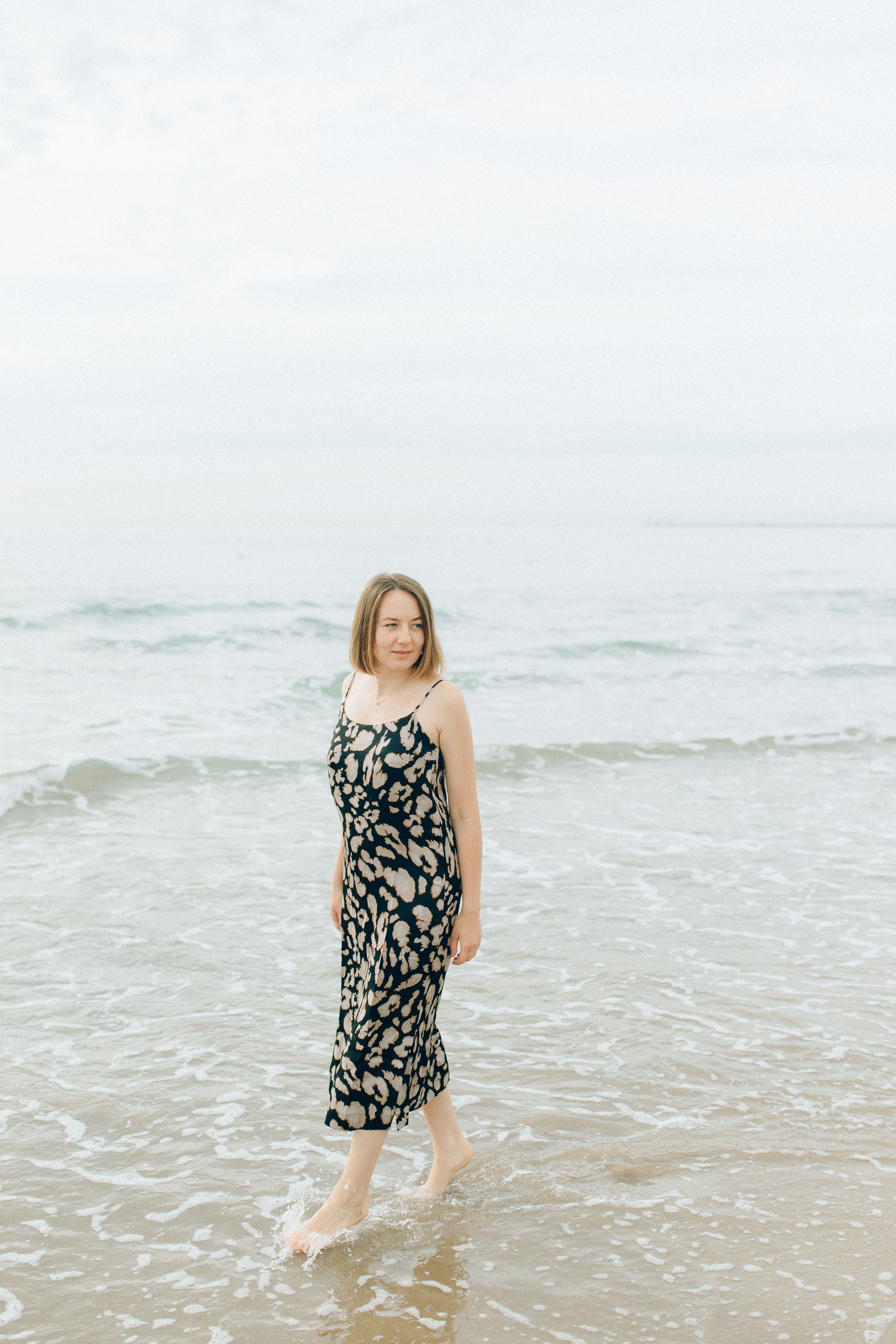 一名赤脚女子穿着连衣裙、留着短发，沿着平静的海滩散步