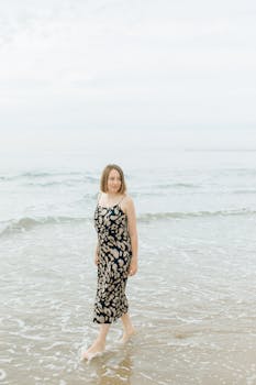 A woman with short hair in a dress walks barefoot along a calm beach shore.