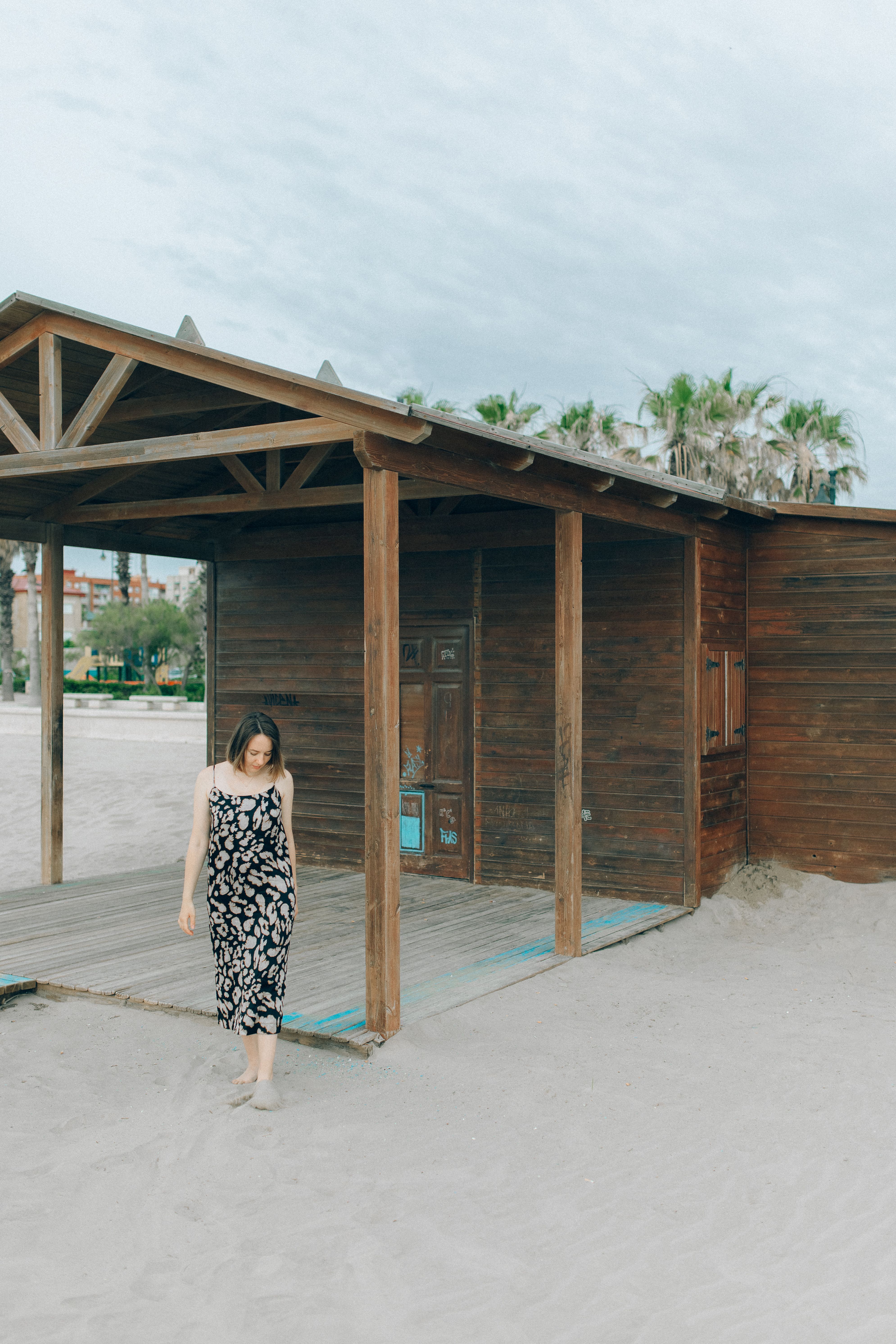 Woman in a floral dress walking by a rustic cabin on a serene beach with sand and palm trees.