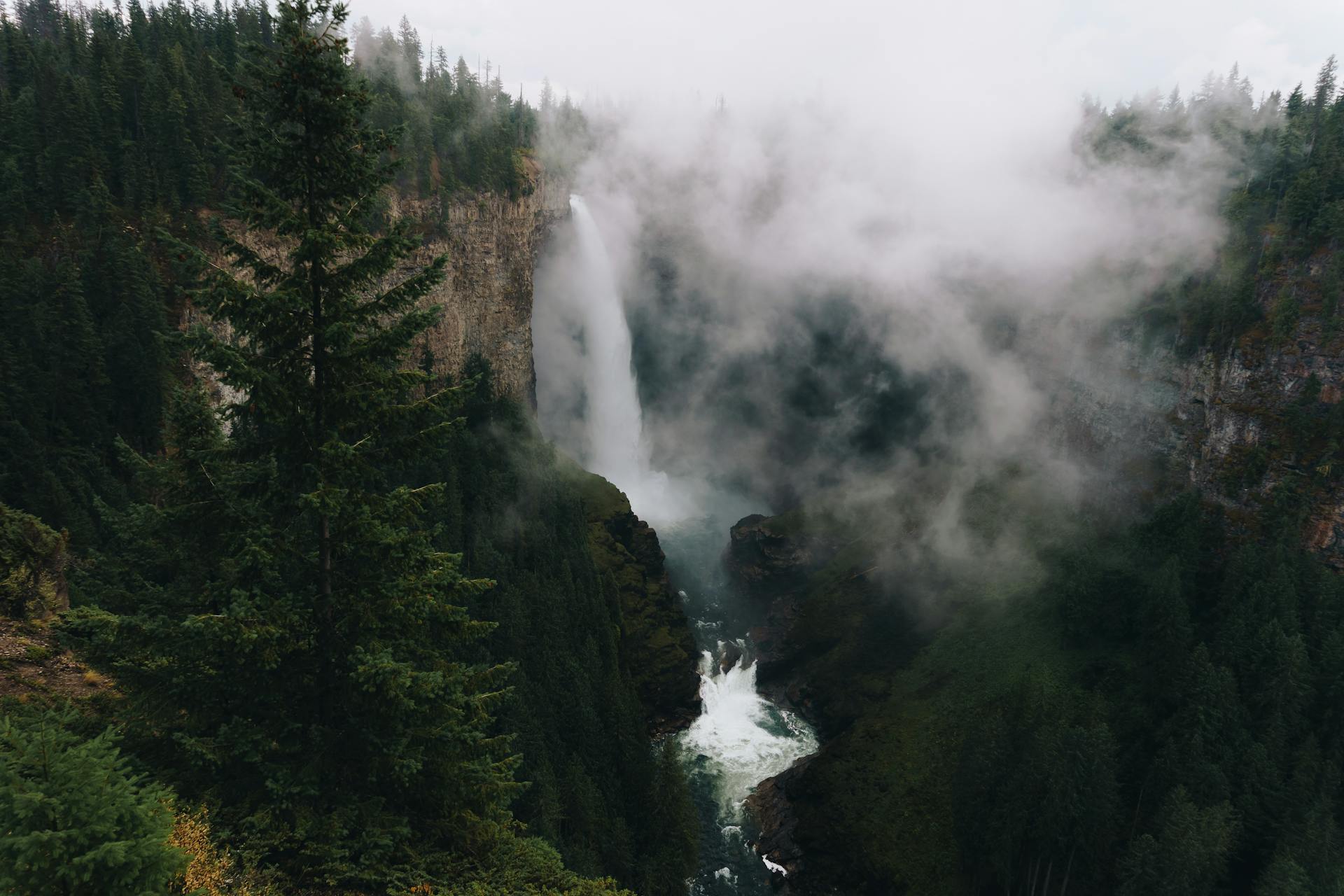 Waterfall in mountainous terrain with evergreen forest against cloudy sky