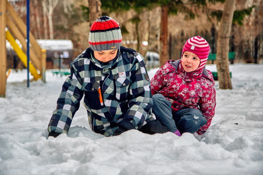 Two children enjoy playing in the snow at a park during winter, wearing colorful warm clothing.