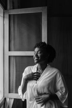 Black and white photo of a woman in a bathrobe smiling by a window, enjoying a tranquil morning.