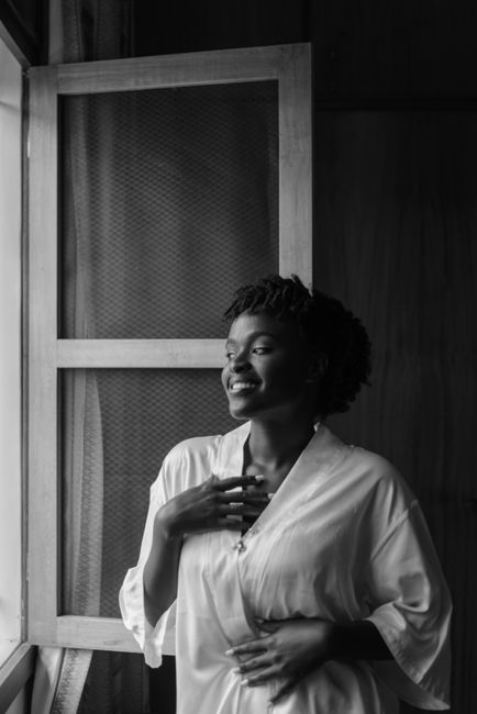Black and white cheerful African American female in white bathrobe standing near window in morning