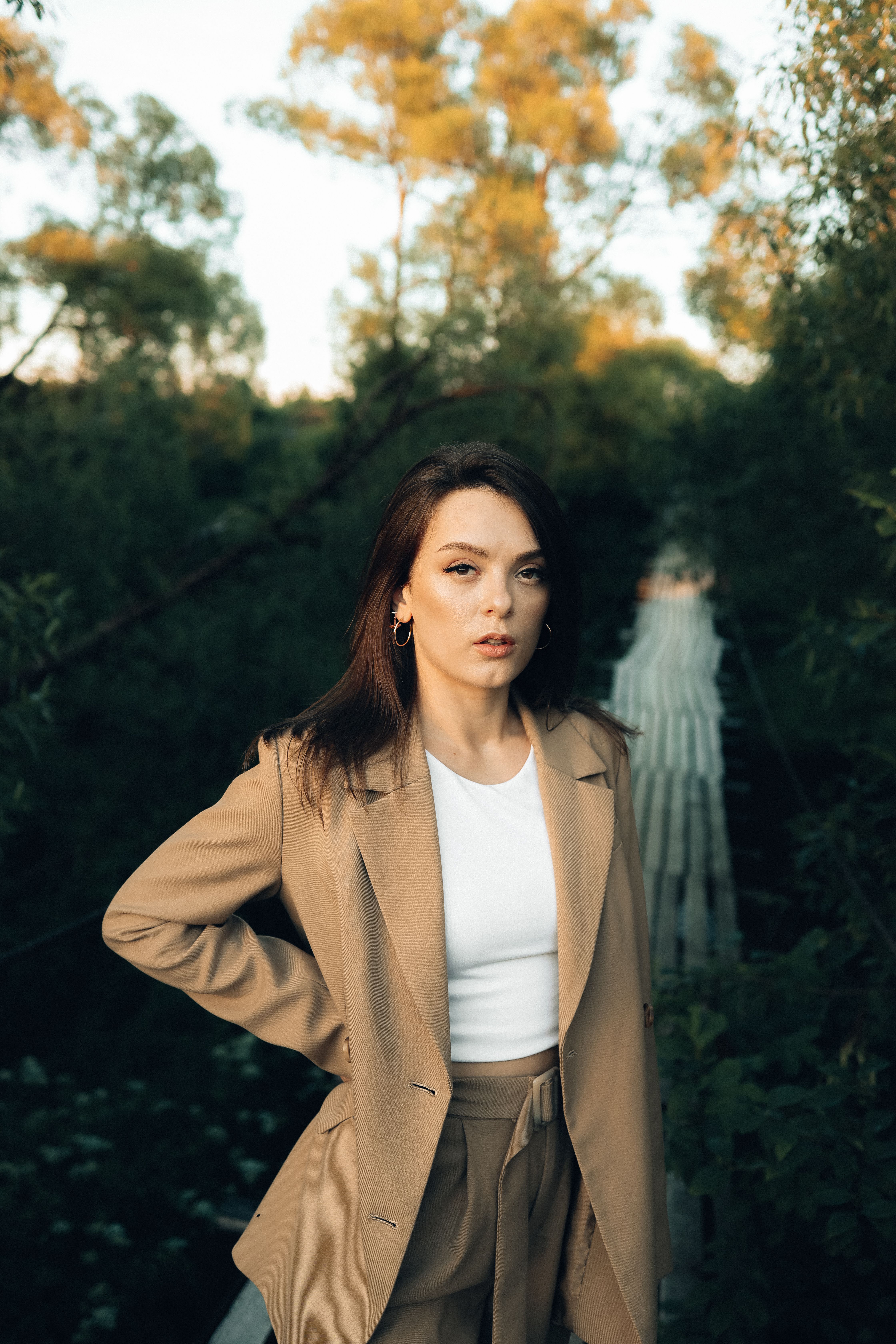 一位穿着棕褐色西装外套的时尚女性，自信地站在一座被大自然环绕的桥上