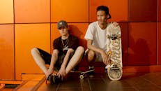 两名年轻的滑冰者拿着滑板，一起靠在明亮的墙上，散发着休闲风格