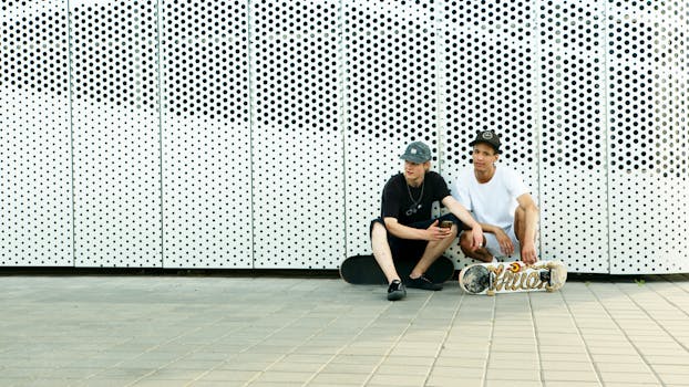 Two male skateboarders sitting against a dotted wall in casual attire, enjoying a break.