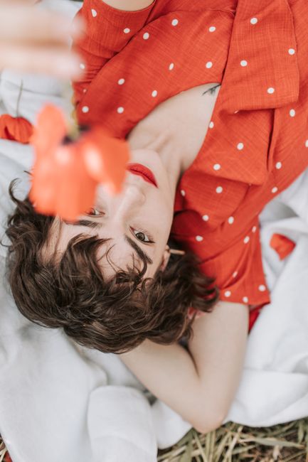 A woman in an orange polka dot dress lies on a white blanket holding a red flower.