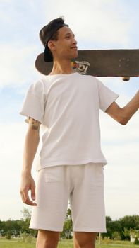 Young male skateboarder holding a skateboard in a sunny park setting.