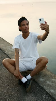 Casual young man in white outfit taking a selfie by a calm water's edge at sunset.