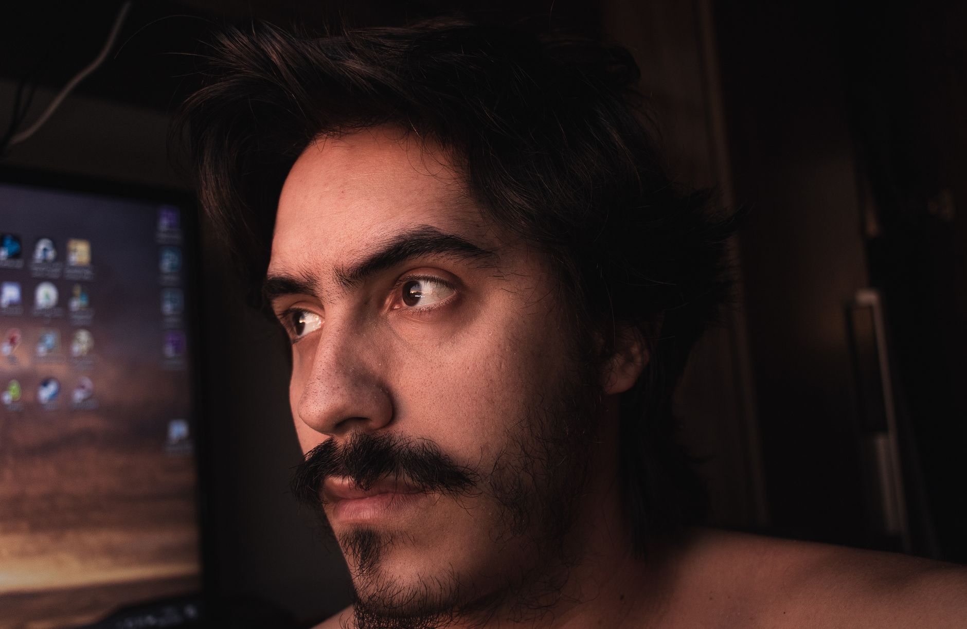 Jovem de bigode e tronco nu numa divisão escura em frente a um ecrã de computador ligado