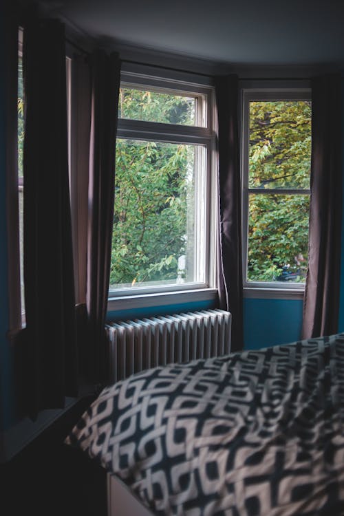 Comfortable bed placed in room in front of window overlooking garden with lush green trees