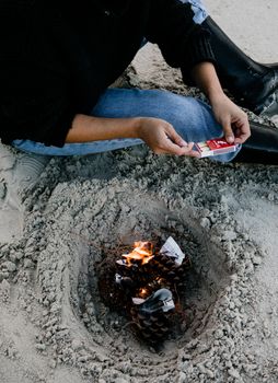 Person using matches to start a fire on a sandy beach with pine cones.