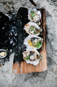Freshly grilled oysters with herbs on a rustic wood plank over beach sand.