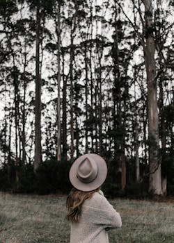 一位戴着帽子的女人若有所思地站着，凝视着那片宁静而茂密的森林