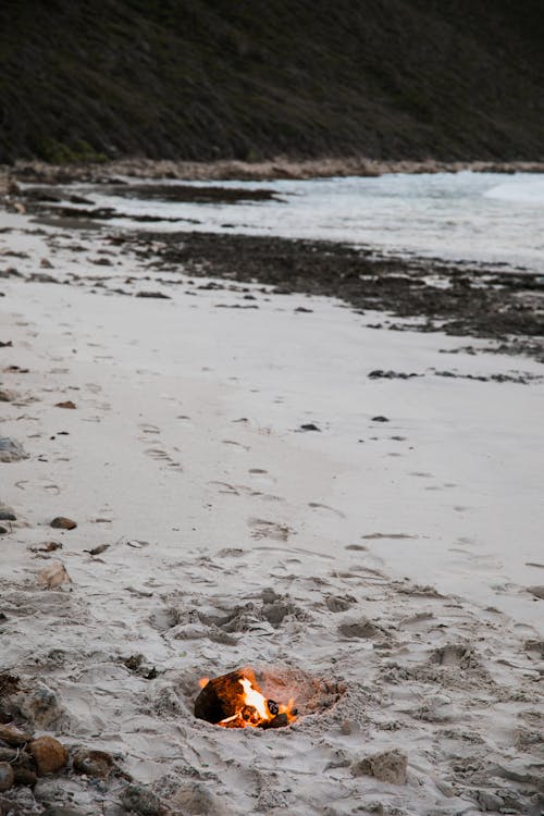 Sandy shore with bonfire and pebbles near foamy sea water splashing on coast