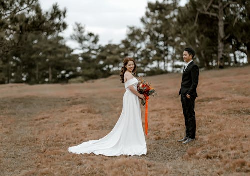 Free Full body of Asian groom in elegant suit standing with hands in pockets near bride in wedding dress with hem and bouquet in hands with green trees on blurred background Stock Photo