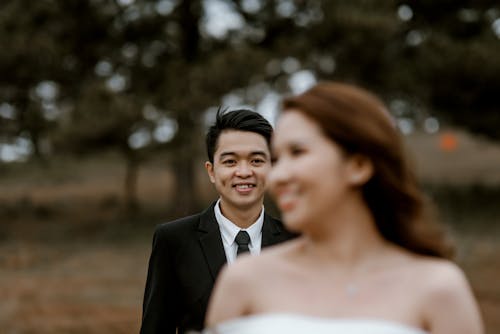 Free Smiling Asian groom standing in field with trees on blurred background behind elegant blurred bride with bare shoulders in wedding ceremony Stock Photo