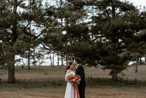 Free Side view of groom in suit kissing bride with flowers on forehead while standing on grassy field near trees during wedding celebration Stock Photo