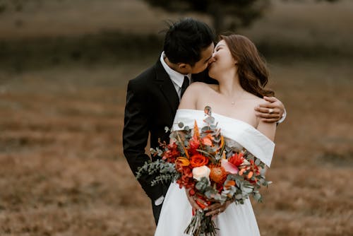 Free Loving Asian groom and bride with bouquet of flowers wearing wedding outfits kissing while standing in nature with blurred background in daylight Stock Photo