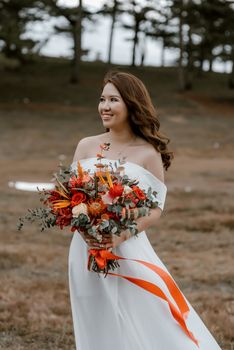 Cheerful young smiling  Asian bride walking with vivid bouquet of flowers and looking away on blurred background of forest